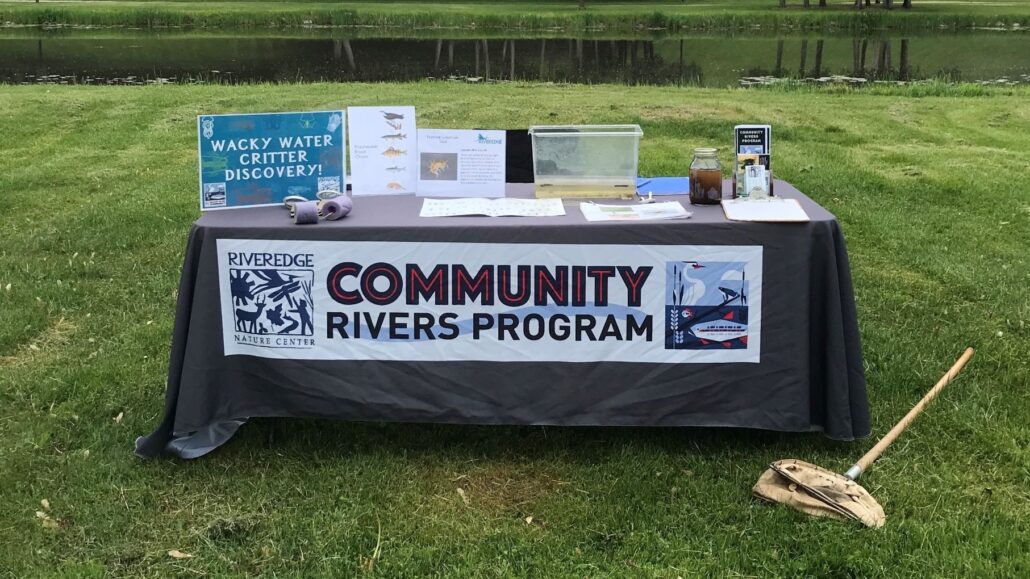 a table with the CRP logo and brochures set up in front of the Milwaukee River on green grass