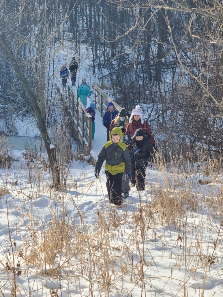 a small group of kids and adults bundled in winter gear hike across a wooden foot bridge through the Riveredge forest into the prairie