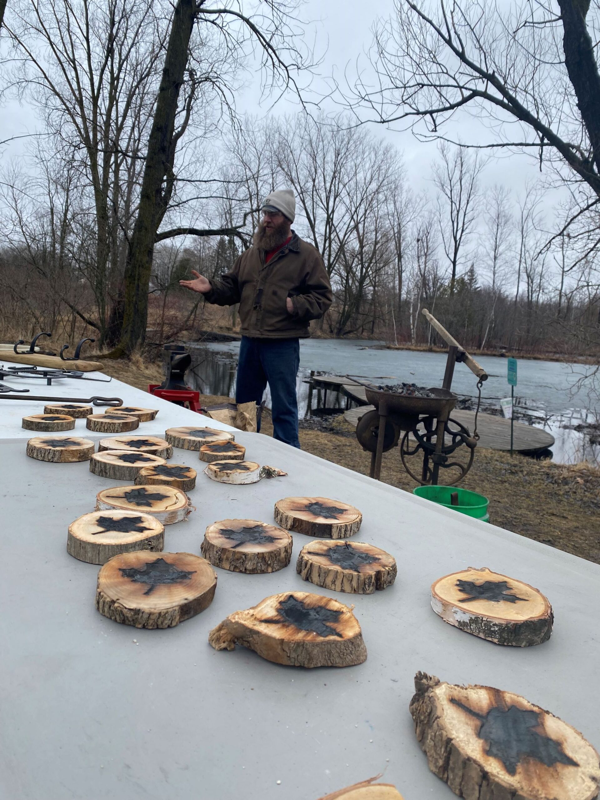 a table with many small tree cookies with maple leaf shapes burned onto them. A blacksmith stands behind the table with trees in the background