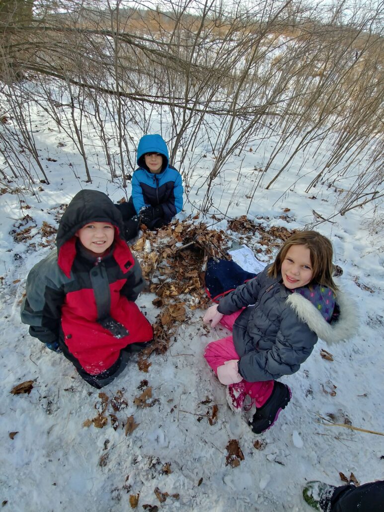 3 kids smile at the camera while sitting around their built animal shelter in winter