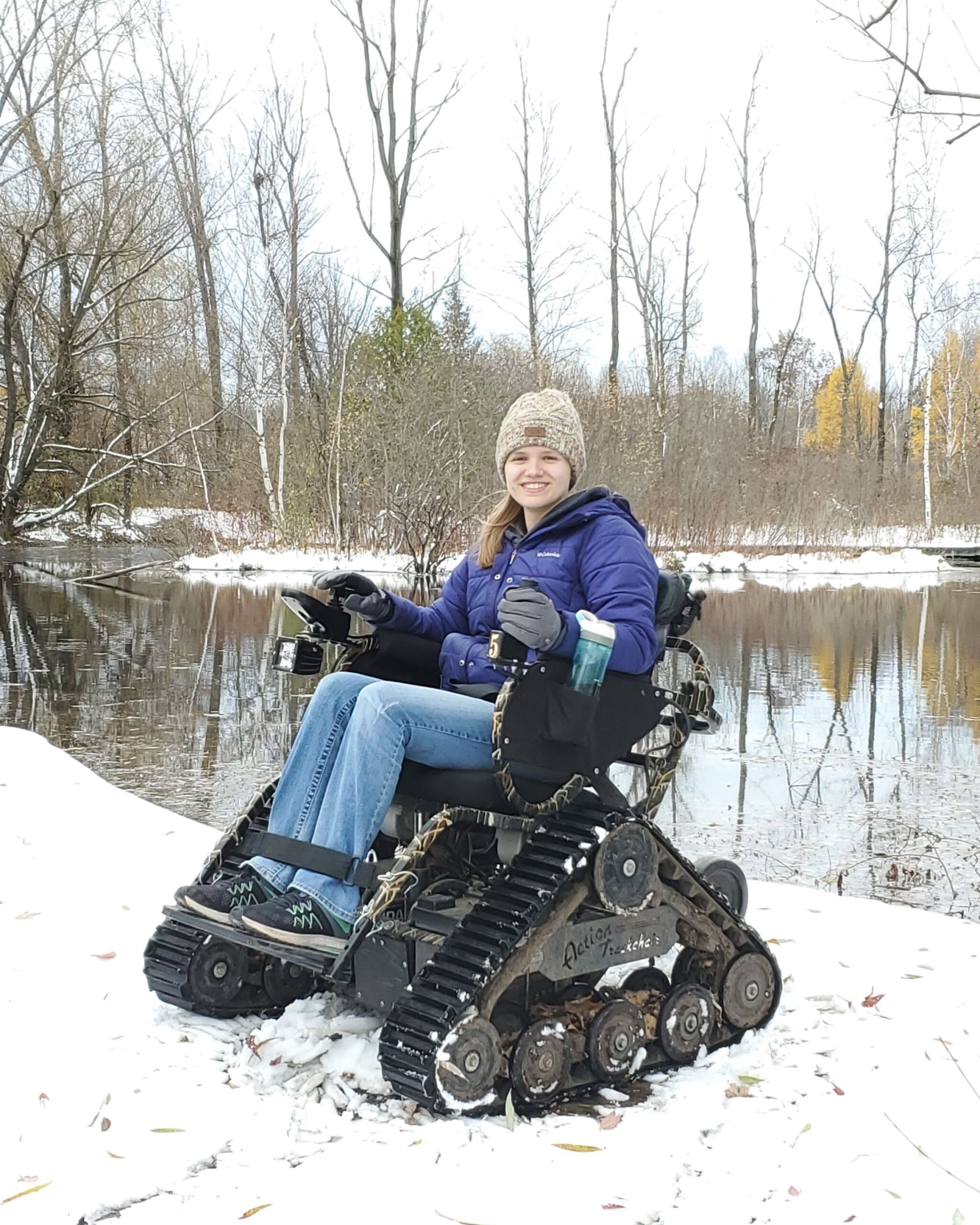 a woman sits in an all terrain wheelchair in front of a Riveredge pond in winter with snow on the ground