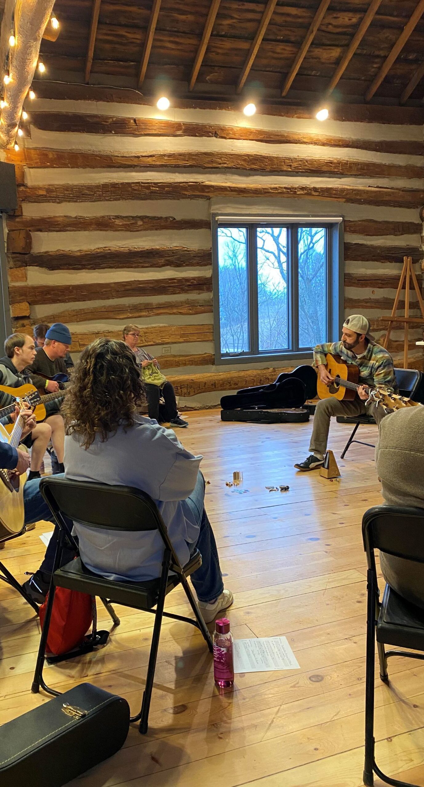 a group of people sit in a circle in the Riveredge Barn playing guitars and watching Luke Cerny play his guitar.