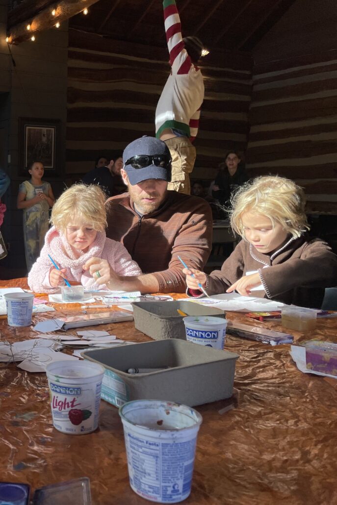 2 young kids sit at a table making crafts with an adult