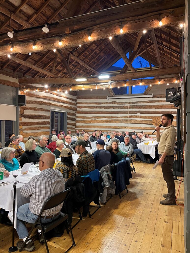 Lots of people sit at tables in the Riveredge Barn listening to John Rakowski speak at the Maple Syrup Supper