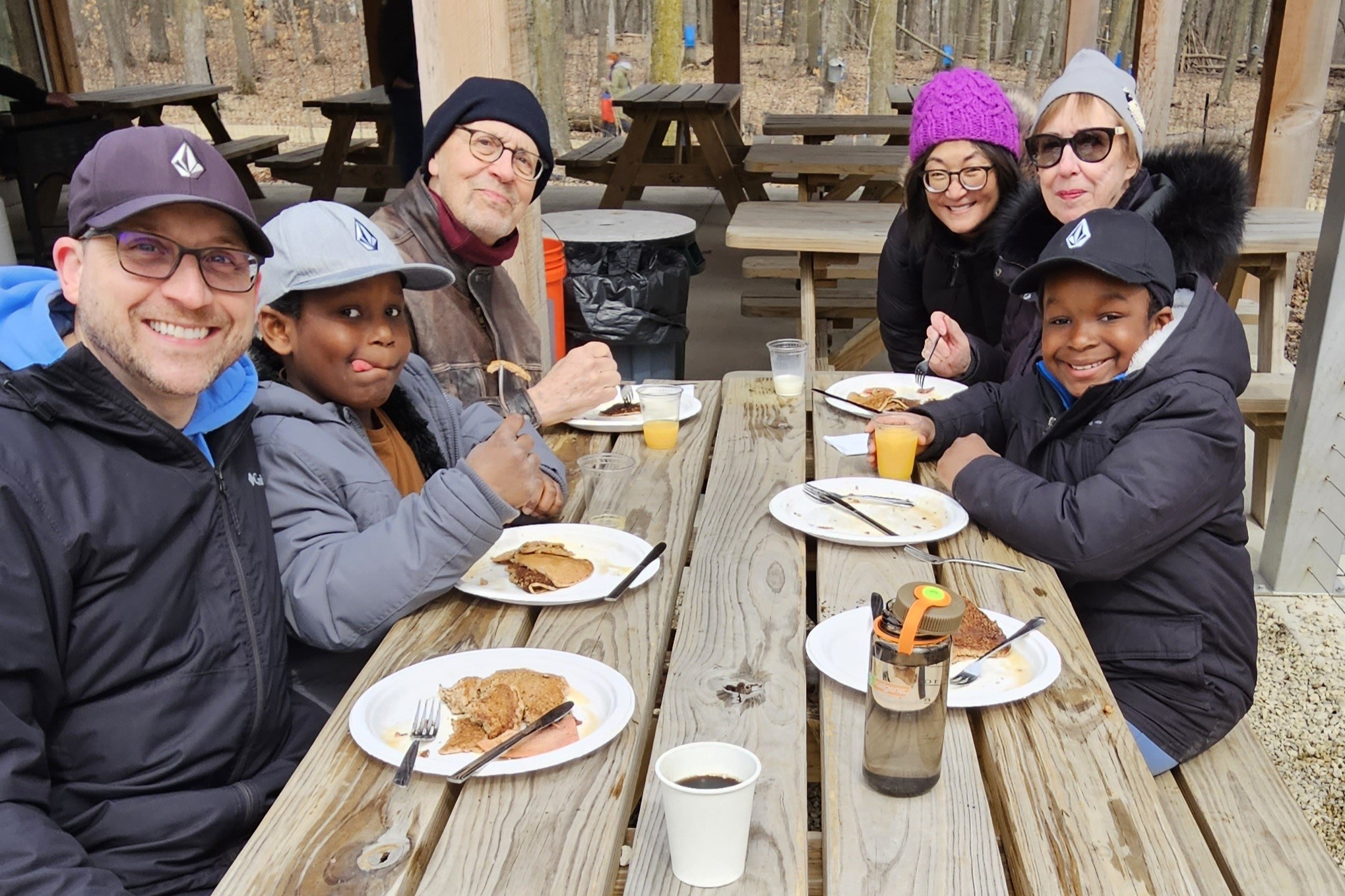 a family sits at a picnic table outside at Riveredge eating pancakes and smiling at the camera