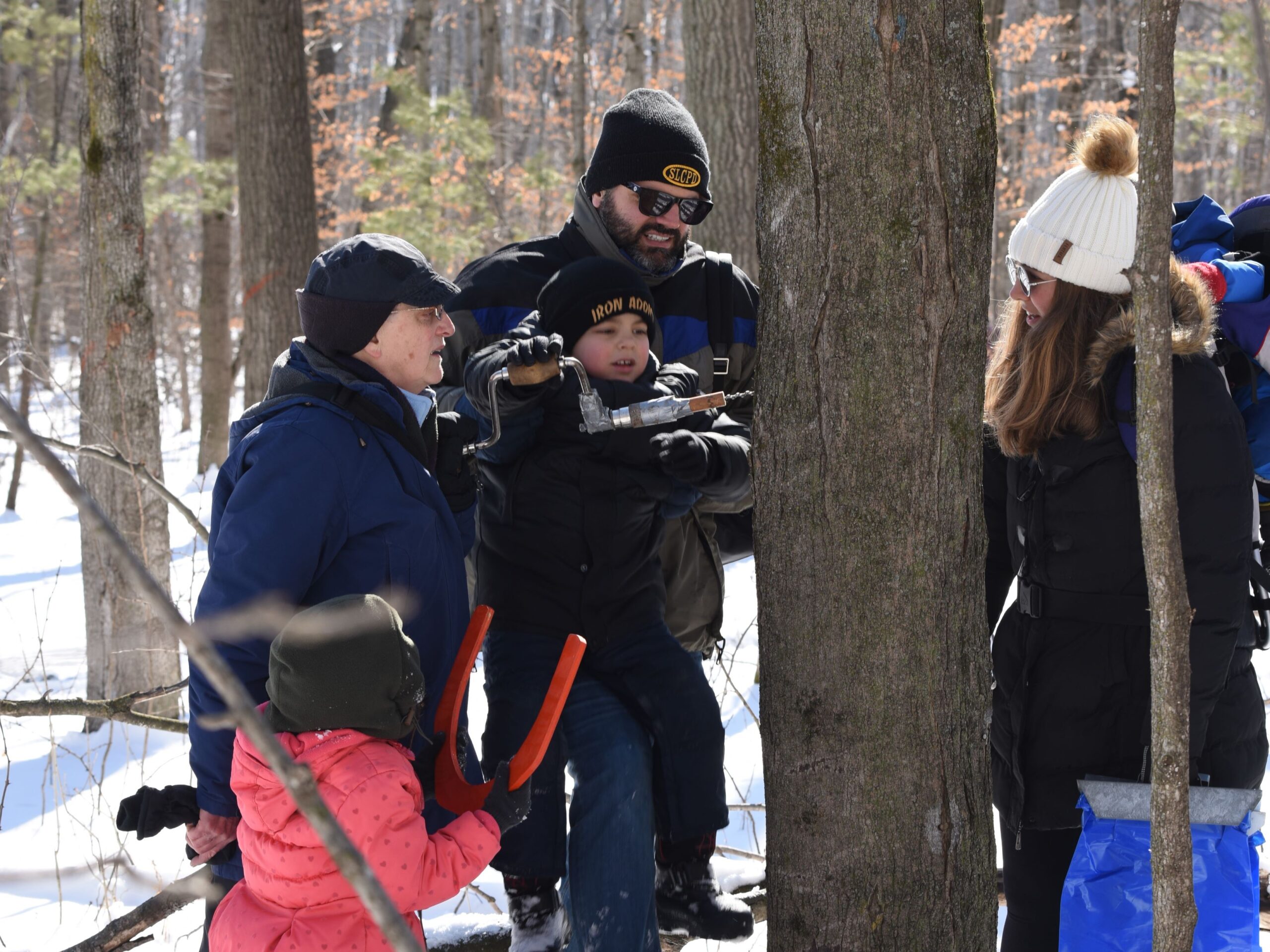 2 young kids and 3 adults tap a maple tree in the snowy Riveredge Sugarbush