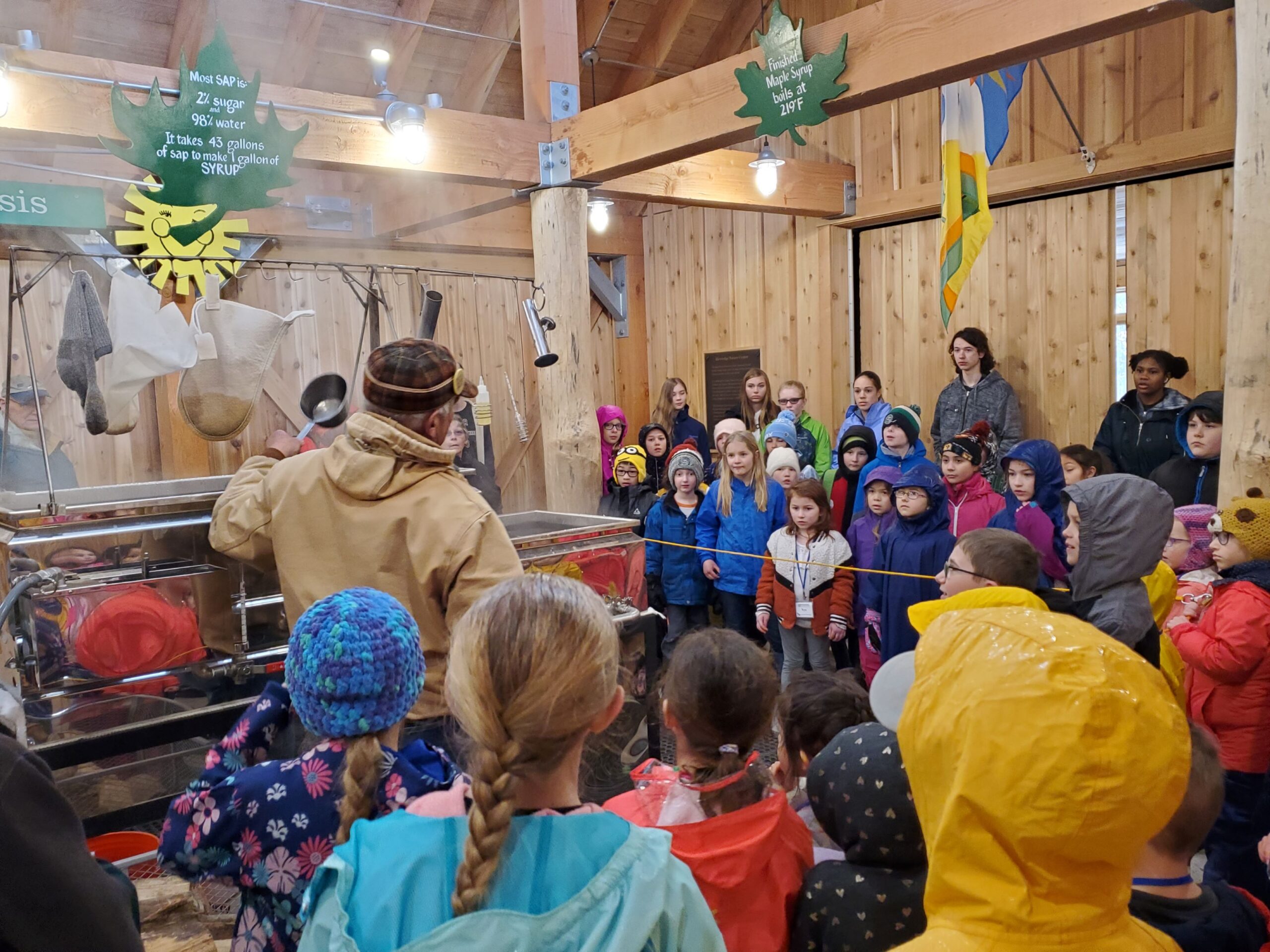 a group of students stands around the steaming sap evaporator in the Riveredge Sugarbush House listening to a program