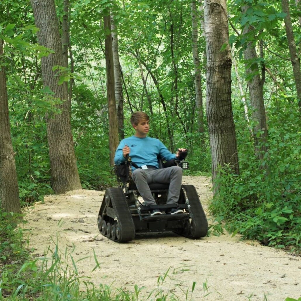 a young man sitting in a trackchair on a gravel trail in a lsuh green Riveredge forest