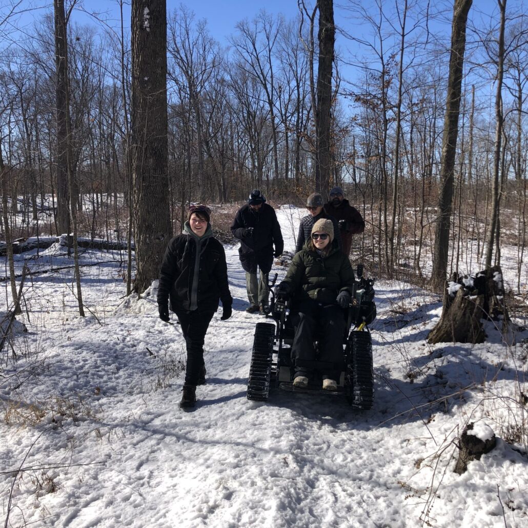 a woman using the trackchair on a snowy winter chair with 3 other adults walking with her