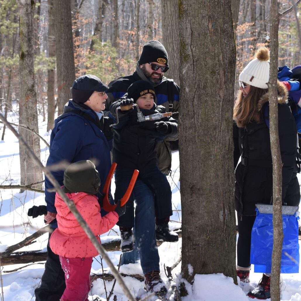 2 young kids and 3 adults tap a maple tree in the snowy Riveredge Sugarbush