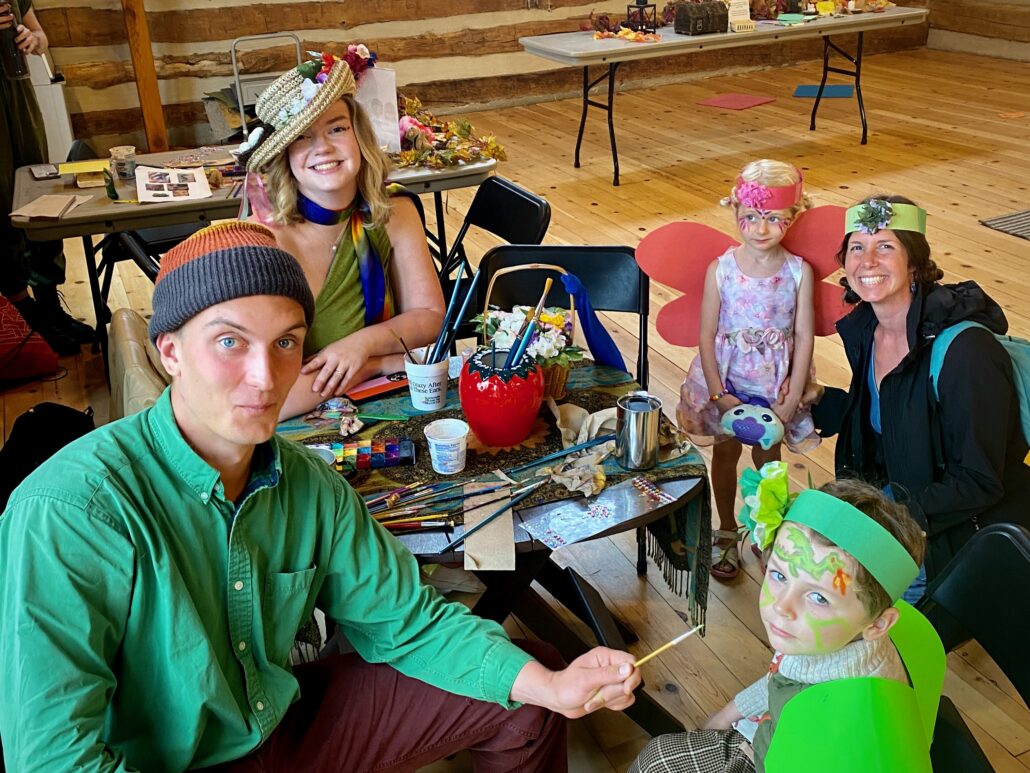 3 adults and 2 kids smiling and sitting around a face painting table at Fairy Fest