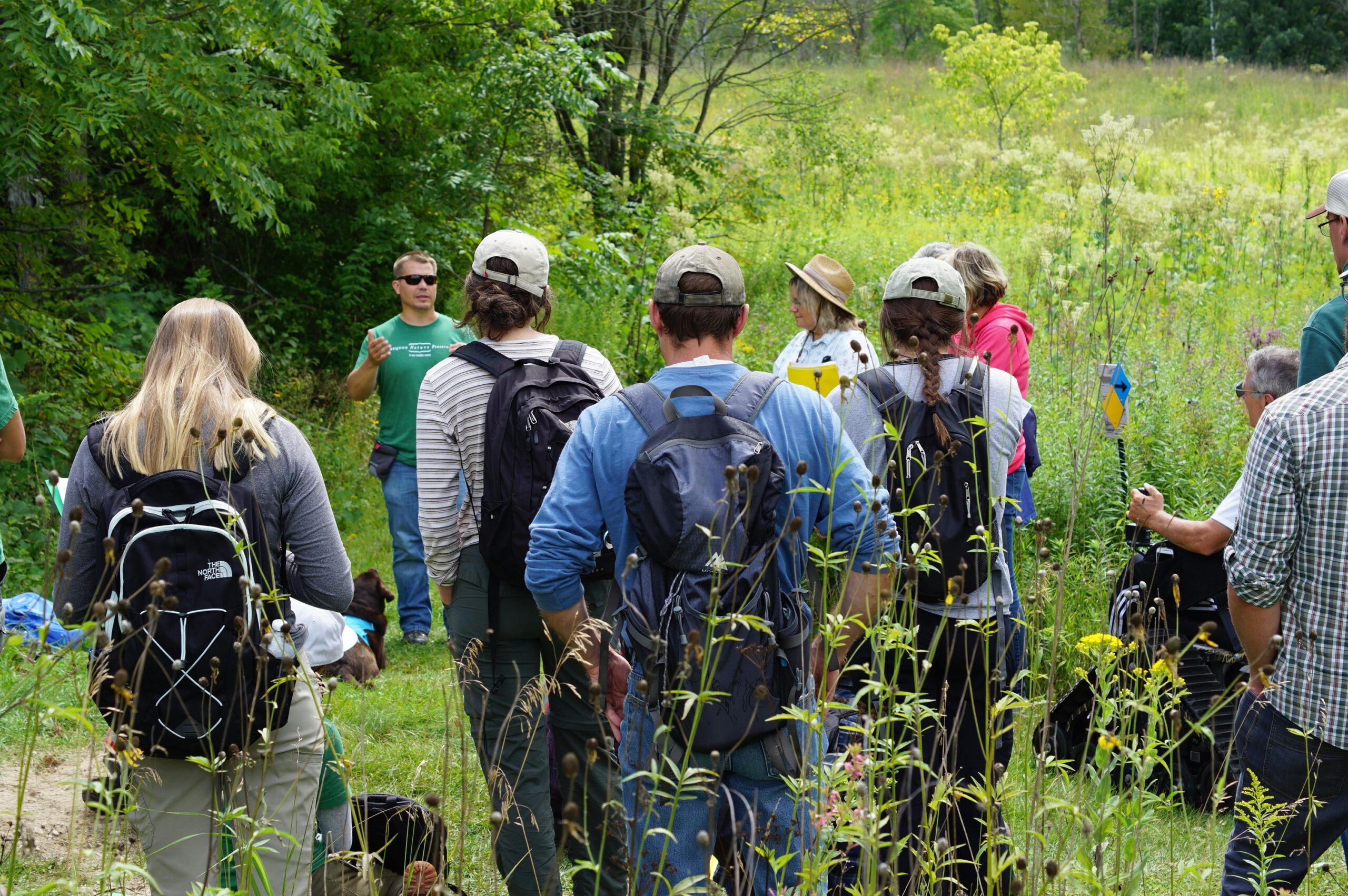 a group of adults standing outside on a sunny summer day and listening to a speaker