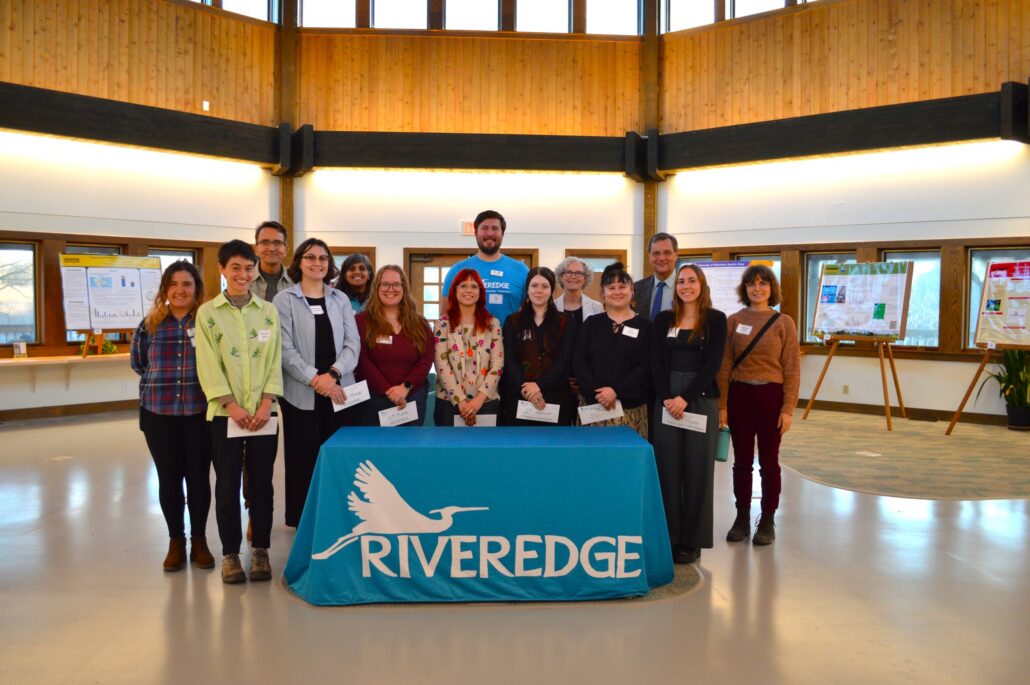 7 student research winners holding their certificates and smiling at the camera along with Riveredge staff