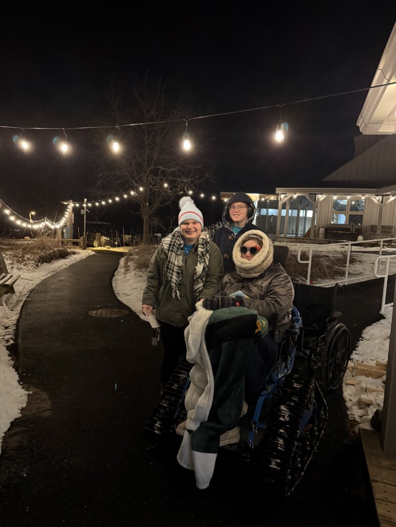 a woman sitting in the Riveredge Trackchair with 2 other adults next to her on a snowy winter evening