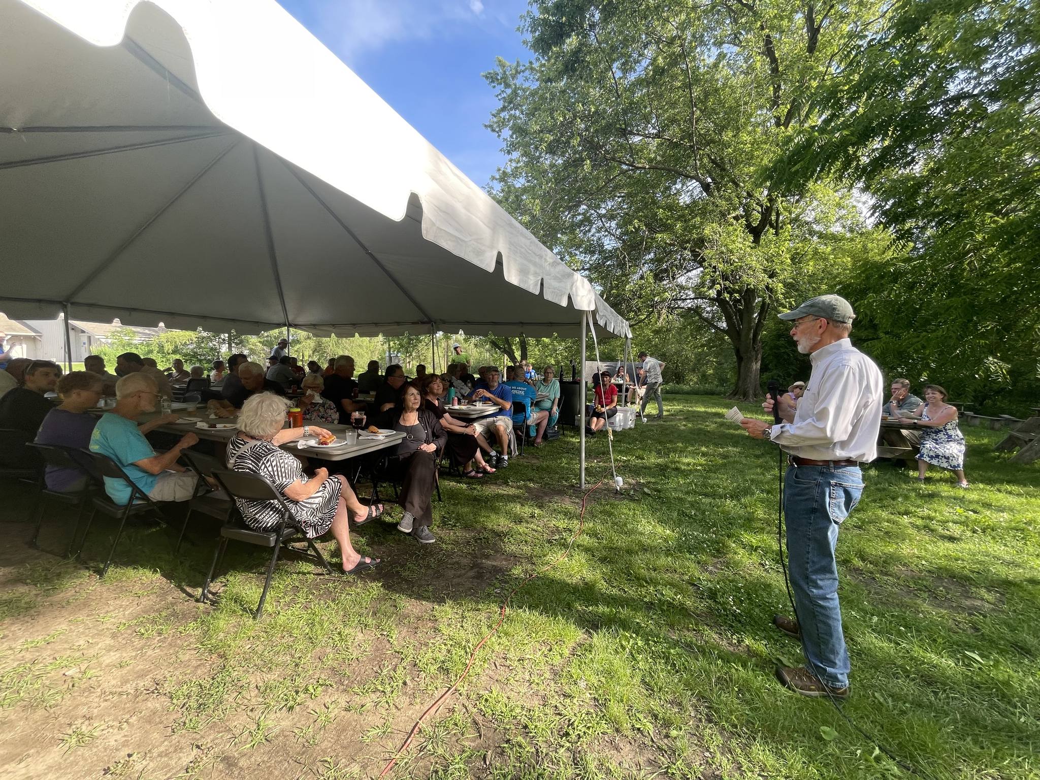 a Riveredge board member speaking into a microphone to a group of Riveredge members and volunteers under a large tent