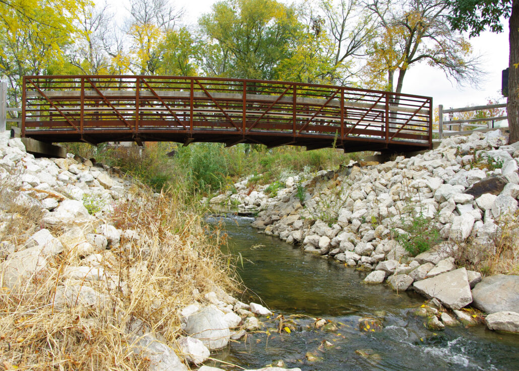 a small stream flowing under a metal foot bridge with rocks along the shoreline